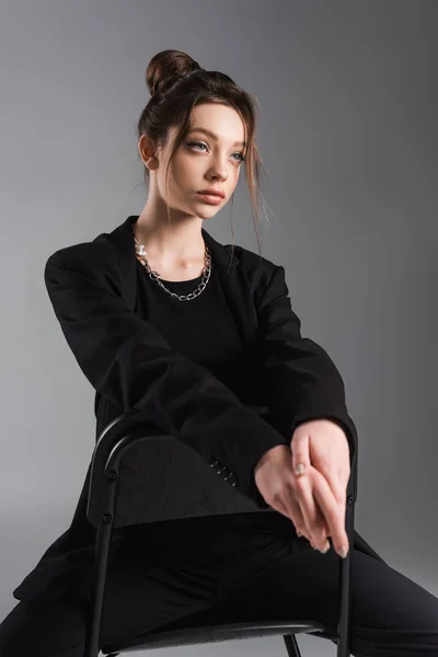 Fashionable woman in black blazer sitting on chair and looking away isolated on grey - foto de stock