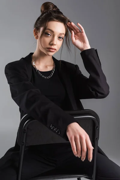 Young woman in black jacket and metal necklace fixing hair while sitting isolated on grey — Photo de stock