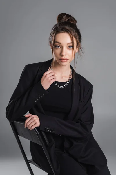 Fashionable woman in black suit and silver necklace looking at camera while sitting isolated on grey - foto de stock