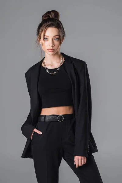 Stylish brunette woman posing with hand in pocket of black trousers isolated on grey — Stockfoto