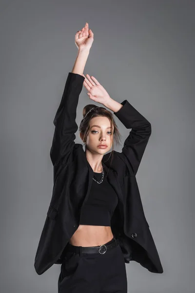 Young woman in black crop top and blazer posing with raised hands isolated on grey — Fotografia de Stock