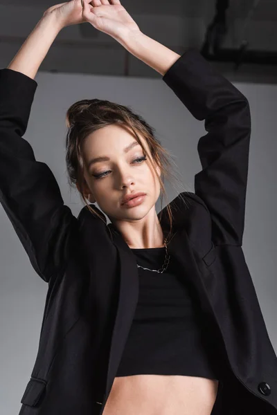 Brunette woman in black jacket posing with arms up on grey background — Stockfoto