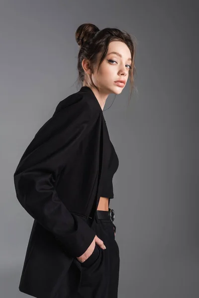 Brunette woman in black blazer posing with hand in pocket isolated on grey — стоковое фото