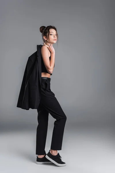 Full length of young woman in black trousers holding jacket and looking at camera on grey background — стоковое фото