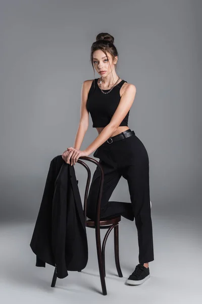 Full length of trendy woman in black clothes posing near jacket on chair on grey background — Stockfoto