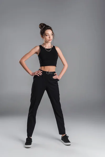 Full length of young woman in black crop top and trousers posing with hands on hips on grey background — Foto stock
