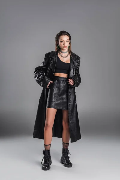 Full length of young woman in black leather coat standing with hand in pocket of skirt on grey background — Photo de stock