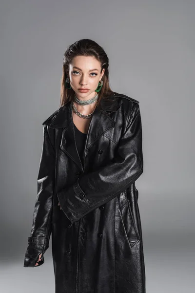 Молода жінка в макіяжі і срібних намистах в чорному шкіряному пальто ізольовані на сірому — стокове фото