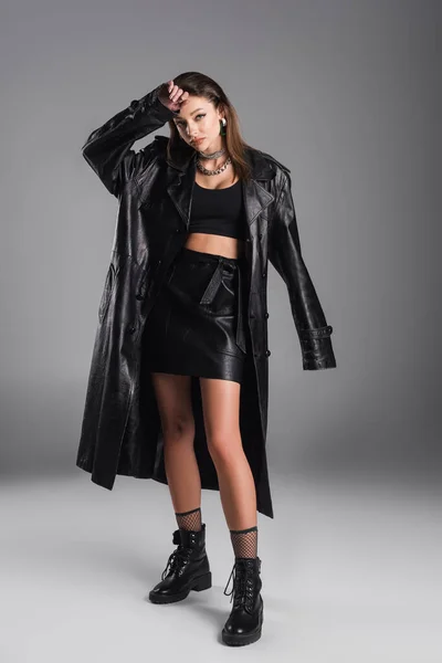 Full length of young woman in black leather coat posing with hand near head on grey background - foto de stock