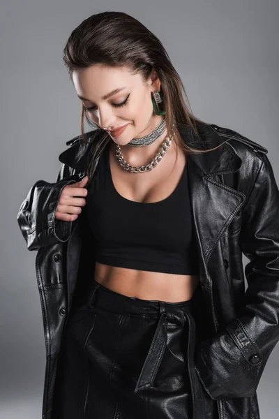 Pretty woman in black leather clothes and silver necklaces smiling isolated on grey - foto de stock