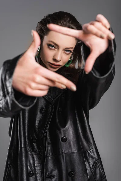 Trendy woman in black leather coat looking at camera and gesturing on blurred foreground isolated on grey — Stock Photo