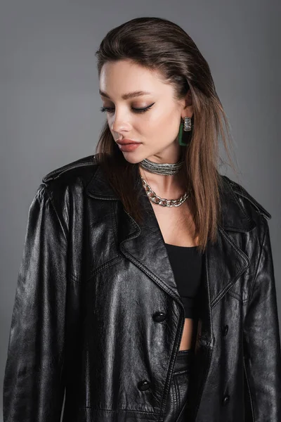 Молода жінка з макіяжем і срібними намистами позує в чорному шкіряному пальто ізольовано на сірому — стокове фото