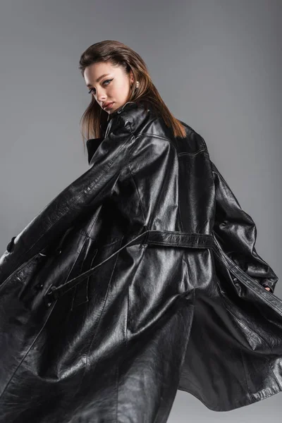 Молода і стильна жінка в чорному шкіряному пальто дивиться на камеру, позує ізольовано на сірому — стокове фото