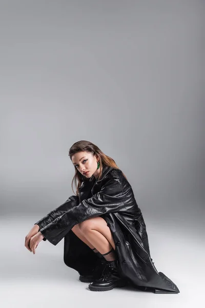 Fashionable brunette woman in black leather outfit sitting on haunches and looking at camera on grey background — Photo de stock