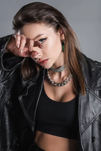 Young woman in black leather coat and silver necklaces obscuring face with hand isolated on grey - foto de stock