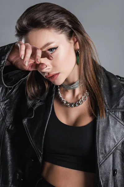 Young woman in black leather coat and silver necklaces obscuring face with hand isolated on grey — Fotografia de Stock