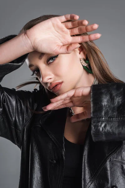 Young and trendy woman in leather clothing posing with hands near face isolated on grey — Stockfoto