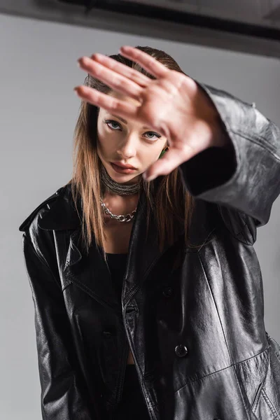 Fashionable woman in black leather coat looking at camera and gesturing on grey background — Fotografia de Stock