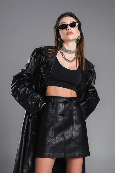 Young woman in dark sunglasses and black leather clothing posing with hands in pockets isolated on grey - foto de stock