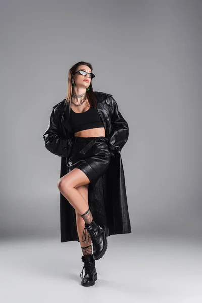 Full length of young woman in black leather clothing and dark sunglasses posing on one leg on grey background - foto de stock