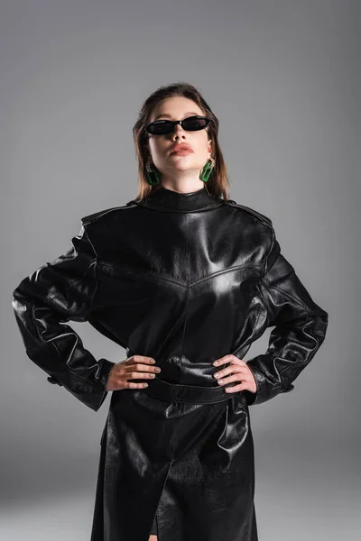 Woman in dark sunglasses wearing leather coat back to front and standing with hands on hips isolated on grey — Fotografia de Stock