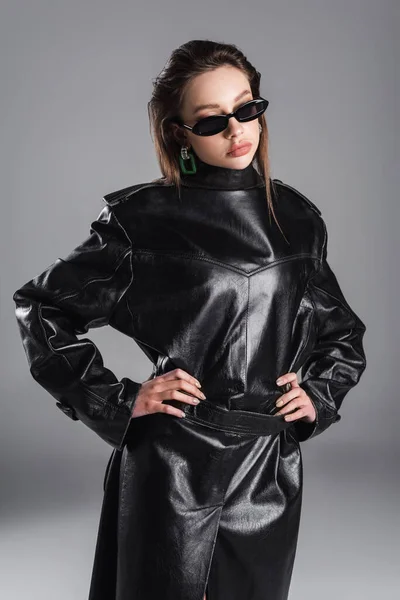 Woman in dark sunglasses posing with hands on hips while wearing leather coat back to front isolated on grey — Fotografia de Stock