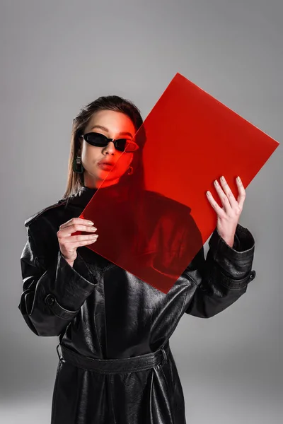 Trendy woman in black leather coat and dark sunglasses posing with red glass isolated on grey - foto de stock