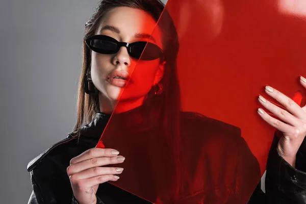 Young brunette woman in black stylish sunglasses posing with red glass isolated on grey — Stockfoto