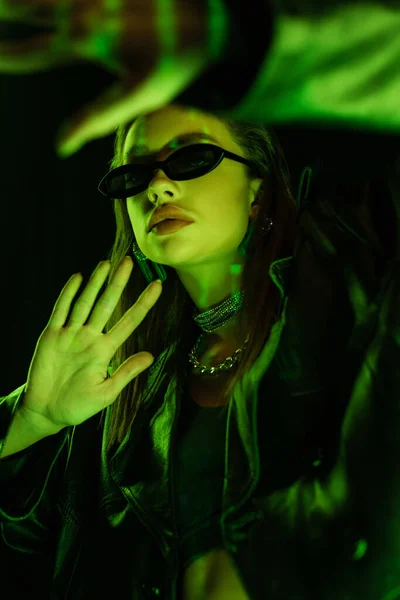 Young woman in dark sunglasses gesturing in green neon light isolated on black — Fotografia de Stock