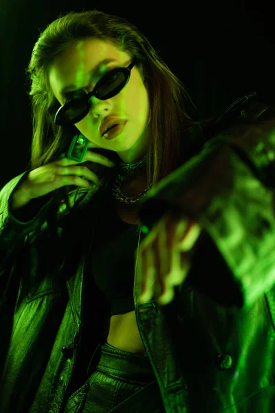 Trendy woman in dark sunglasses and leather coat gesturing in green light isolated on black — Foto stock