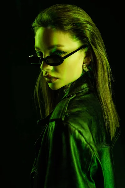 Young brunette woman in dark and trendy sunglasses looking at camera in green light isolated on black — Foto stock