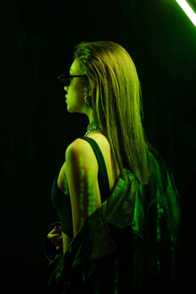 Stylish woman in sunglasses posing with leather coat in green neon light on black background — Fotografia de Stock