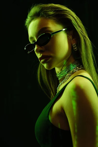 Portrait of stylish woman in dark sunglasses and silver necklaces in green light isolated on black — Stock Photo