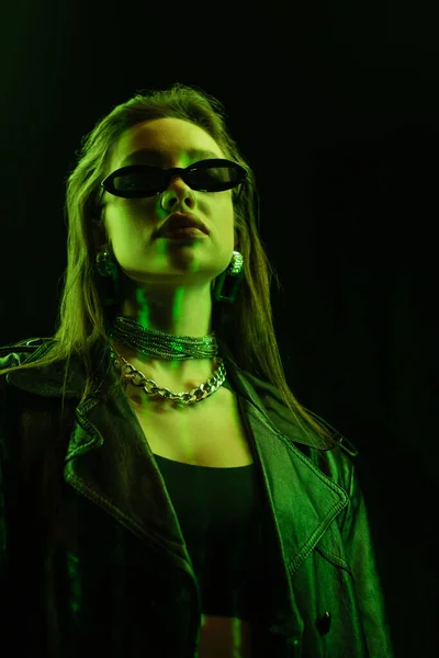 Low angle view of young woman in trendy sunglasses and metal necklaces in green neon light isolated on black — Fotografia de Stock