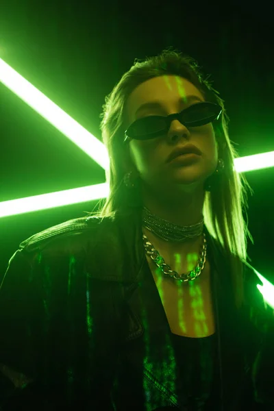 Low angle view of woman in dark sunglasses on black background with green neon light — стоковое фото