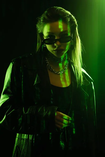 Fashionable woman in sunglasses and leather coat looking at camera on black background with green light - foto de stock