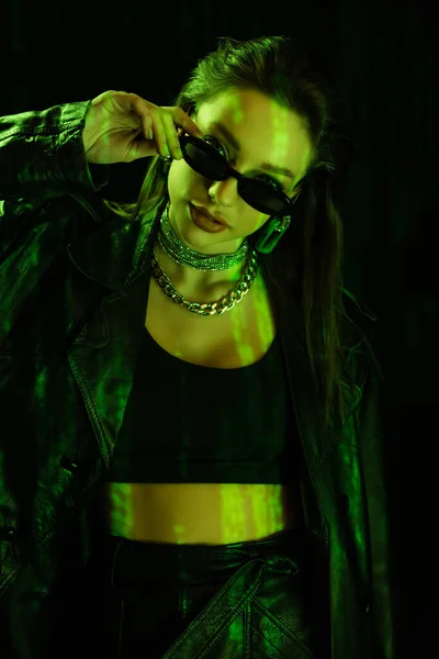 Trendy woman in crop top holding sunglasses and looking at camera in green light isolated on black — стоковое фото