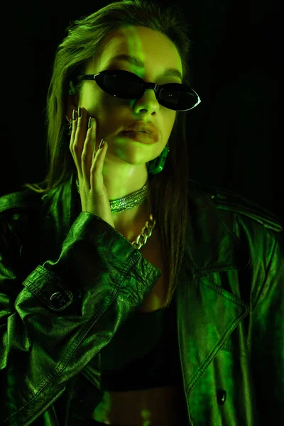 Woman in leather coat and fashionable sunglasses touching face in green light isolated on black — Stock Photo