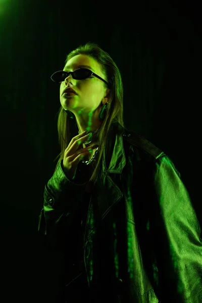 Young woman in leather coat and sunglasses touching neck in green light isolated on black — стоковое фото