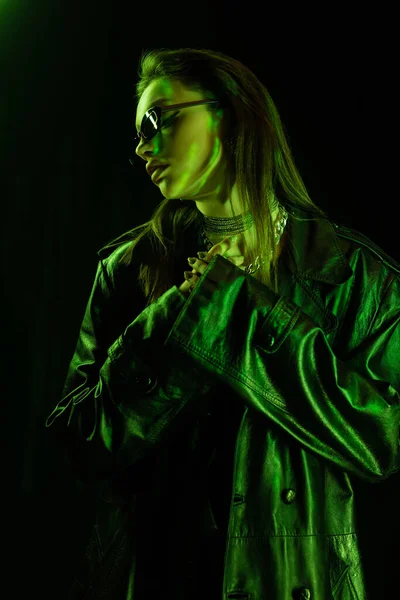 Trendy woman in leather coat and dark sunglasses holding hands near chest in green light isolated on black — Stockfoto