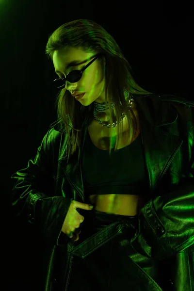 Trendy woman in sunglasses and crop top standing with hand on hip in green light isolated on black — стоковое фото