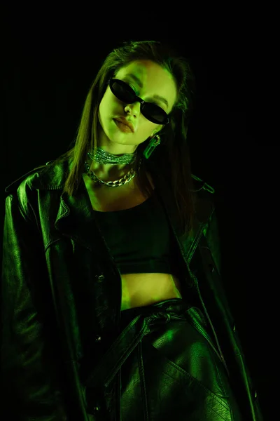 Fashionable woman in leather clothes and sunglasses in green neon light isolated on black — стоковое фото