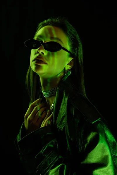 Portrait of brunette woman in trendy sunglasses and silver necklaces in green light isolated on black — Foto stock