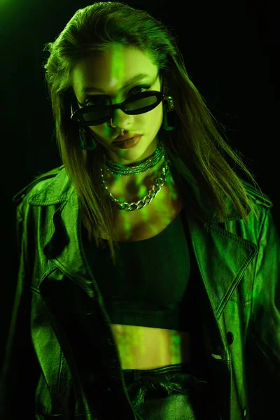 Stylish woman in crop top and dark sunglasses looking at camera in green light isolated on black — Photo de stock