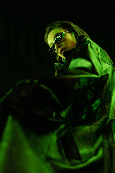 Low angle view of woman in leather coat and dark sunglasses in green light isolated on black — стоковое фото