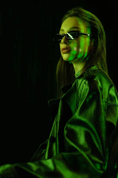 Young brunette woman in leather coat and sunglasses in green neon light isolated on black - foto de stock