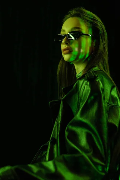 Young brunette woman in leather coat and sunglasses in green neon light isolated on black — стоковое фото