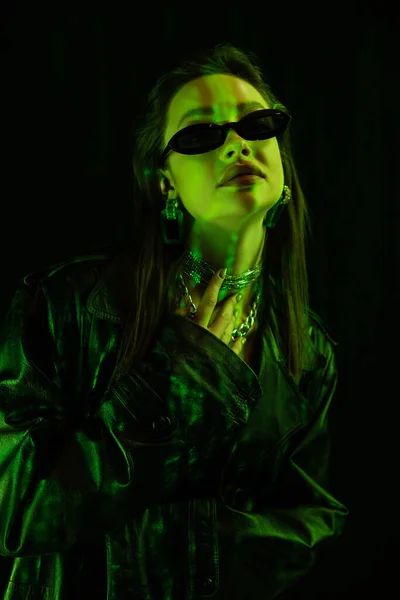 Stylish woman in leather coat and sunglasses touching silver necklaces in green light isolated on black — Stockfoto