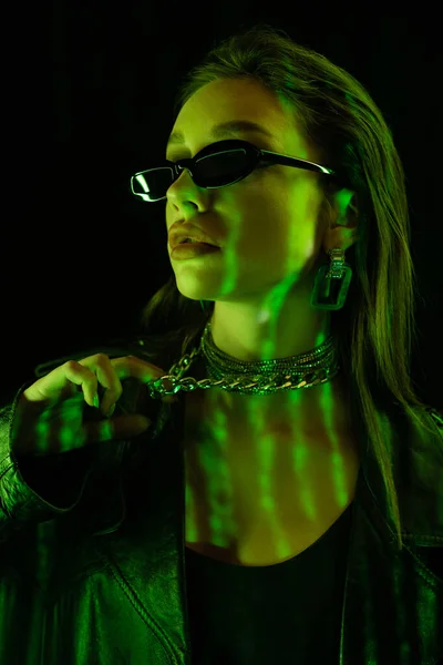 Portrait of young woman in dark sunglasses touching silver necklace in green light isolated on black — Foto stock