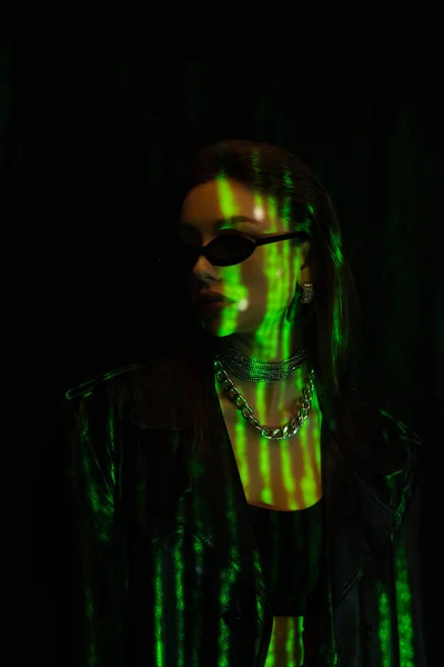 Модна жінка в срібних намистах і стильних сонцезахисних окулярах в зеленому світлі ізольована на чорному — стокове фото